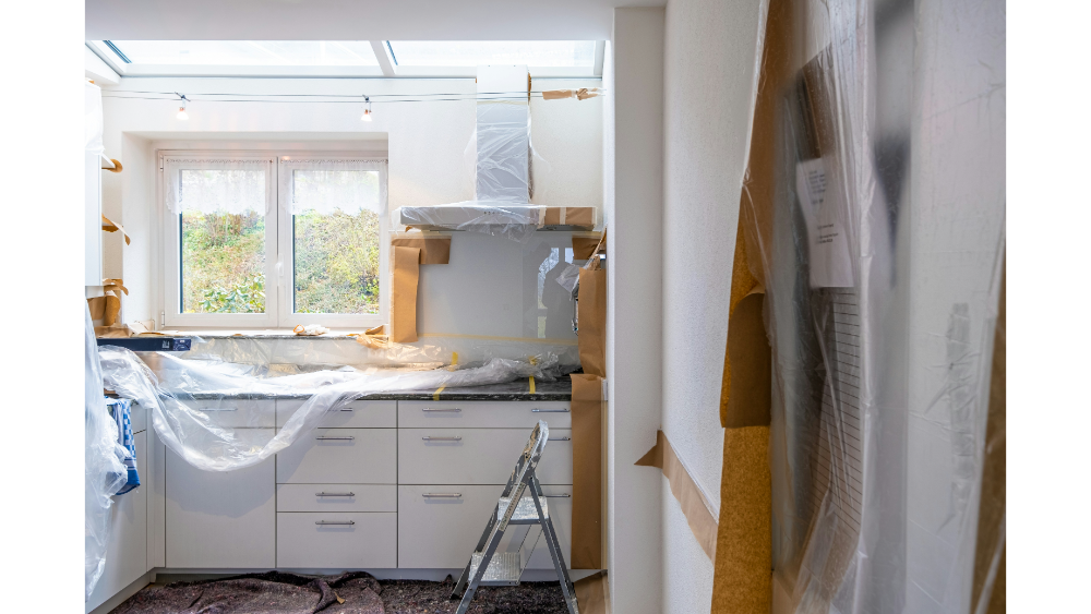 Kitchen Under Renovation With Protective Plastic Coverings, Taped Cabinet Edges, And A Ladder In Front Of A Window With Natural Daylight.